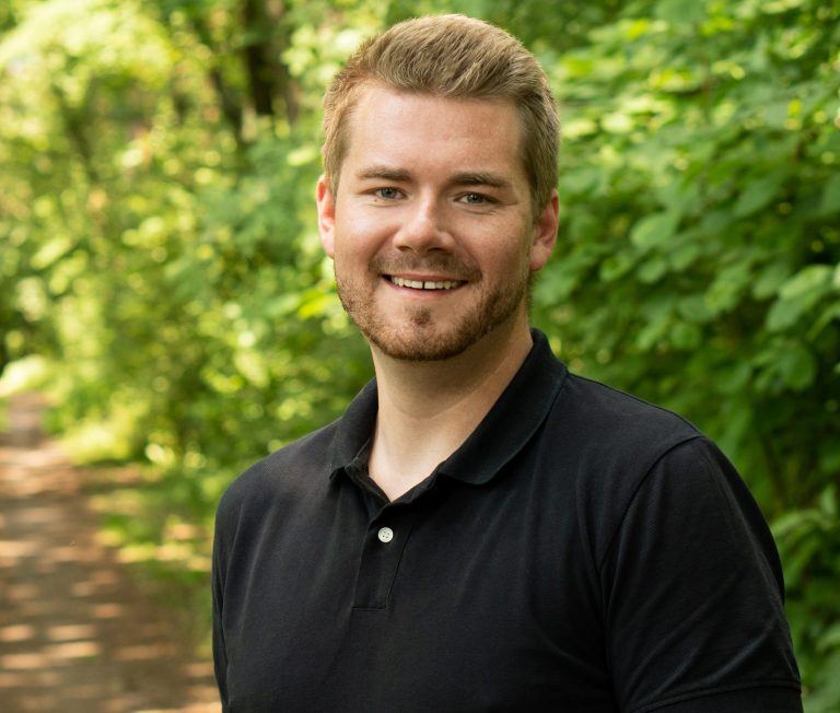 Lächelnder Mann mit blonden Haaren, stehend vor einer grünen Hintergrundnatur.